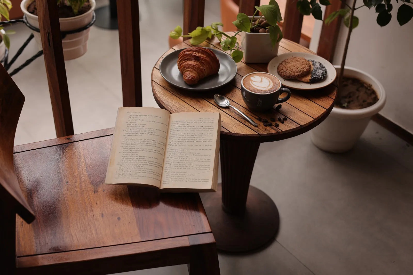 A small wooden cafe table with a cappuccino, a croissant, a cookie and an open book