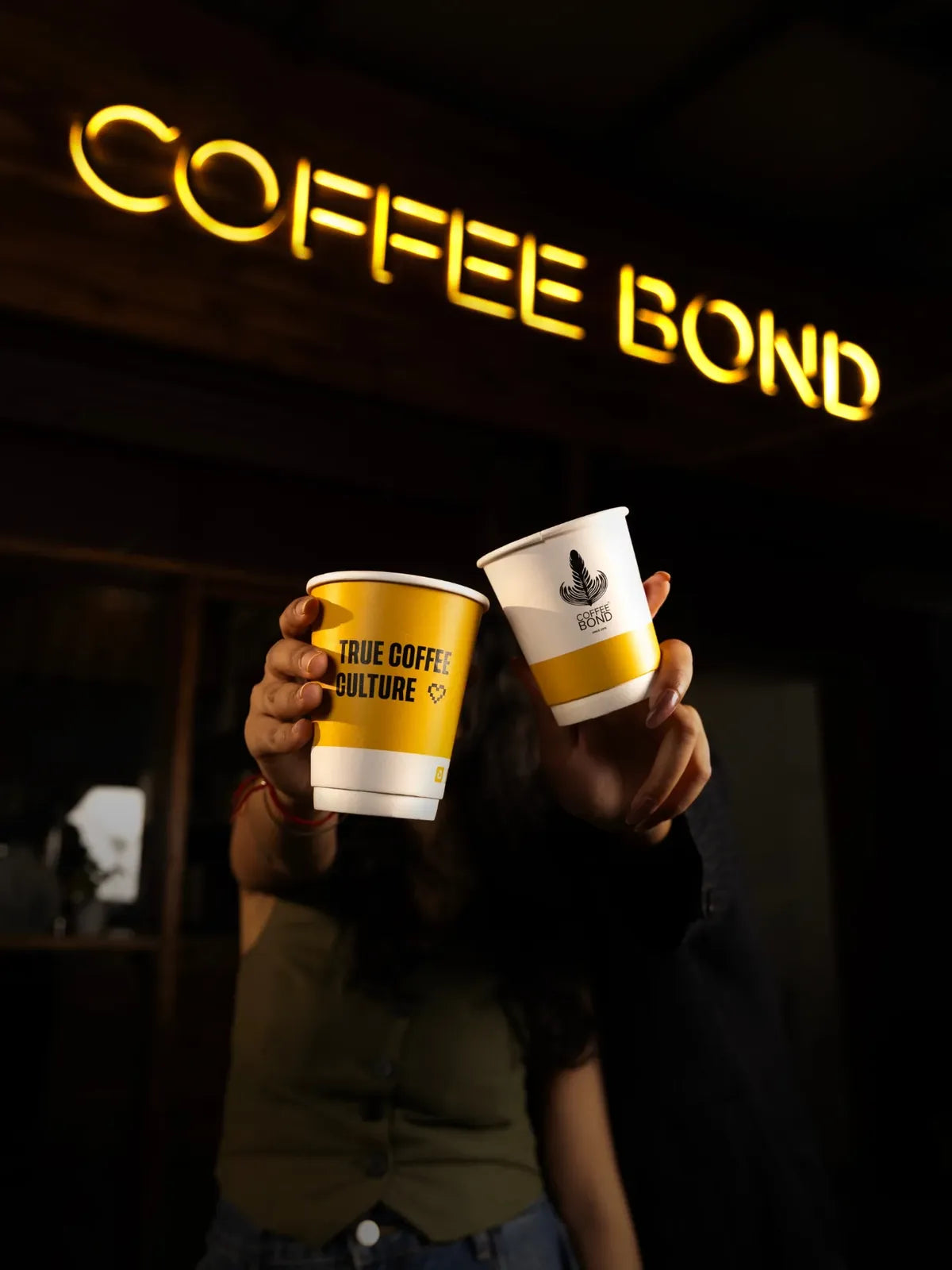 Two hands hold up Coffee Bond paper cups beneath the lit yellow neon Coffee Bond sign