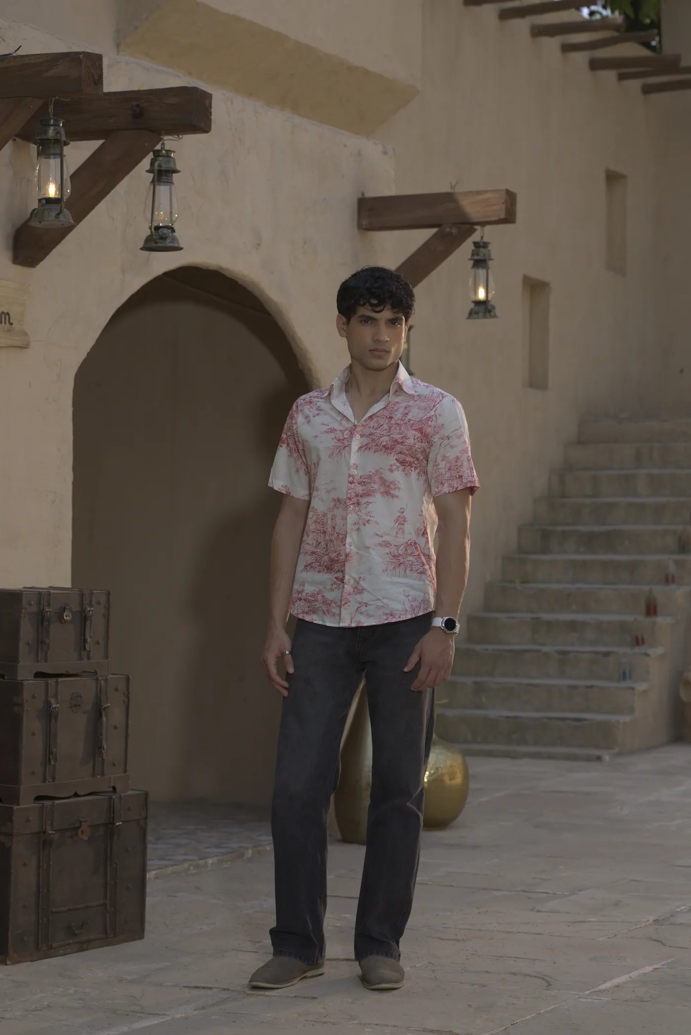 Full-length view of the model in the red toile shirt and dark jeans, in front of an arched doorway and wooden trunks