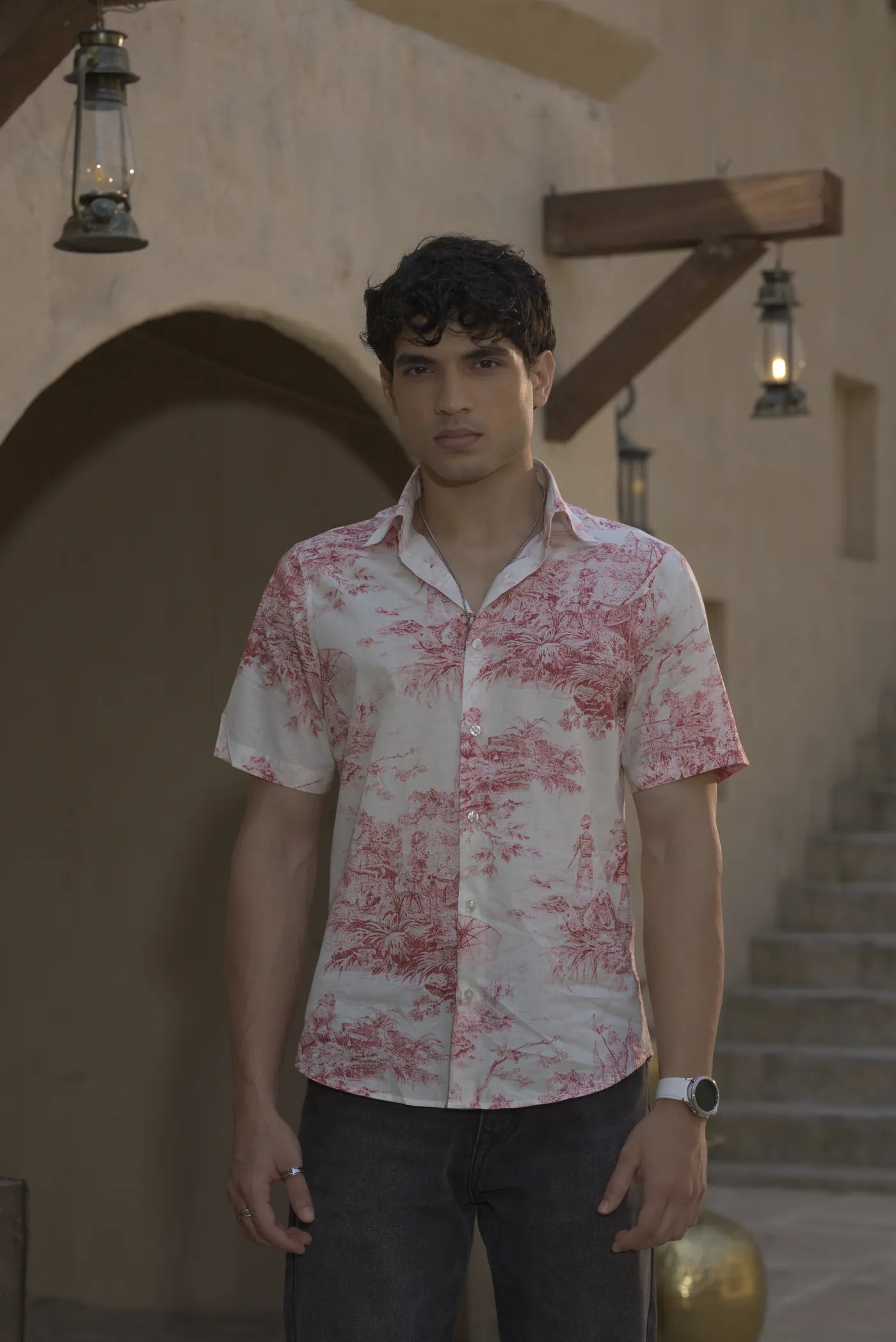 A model in a short-sleeve shirt with a red toile print, facing the camera in a sandstone courtyard