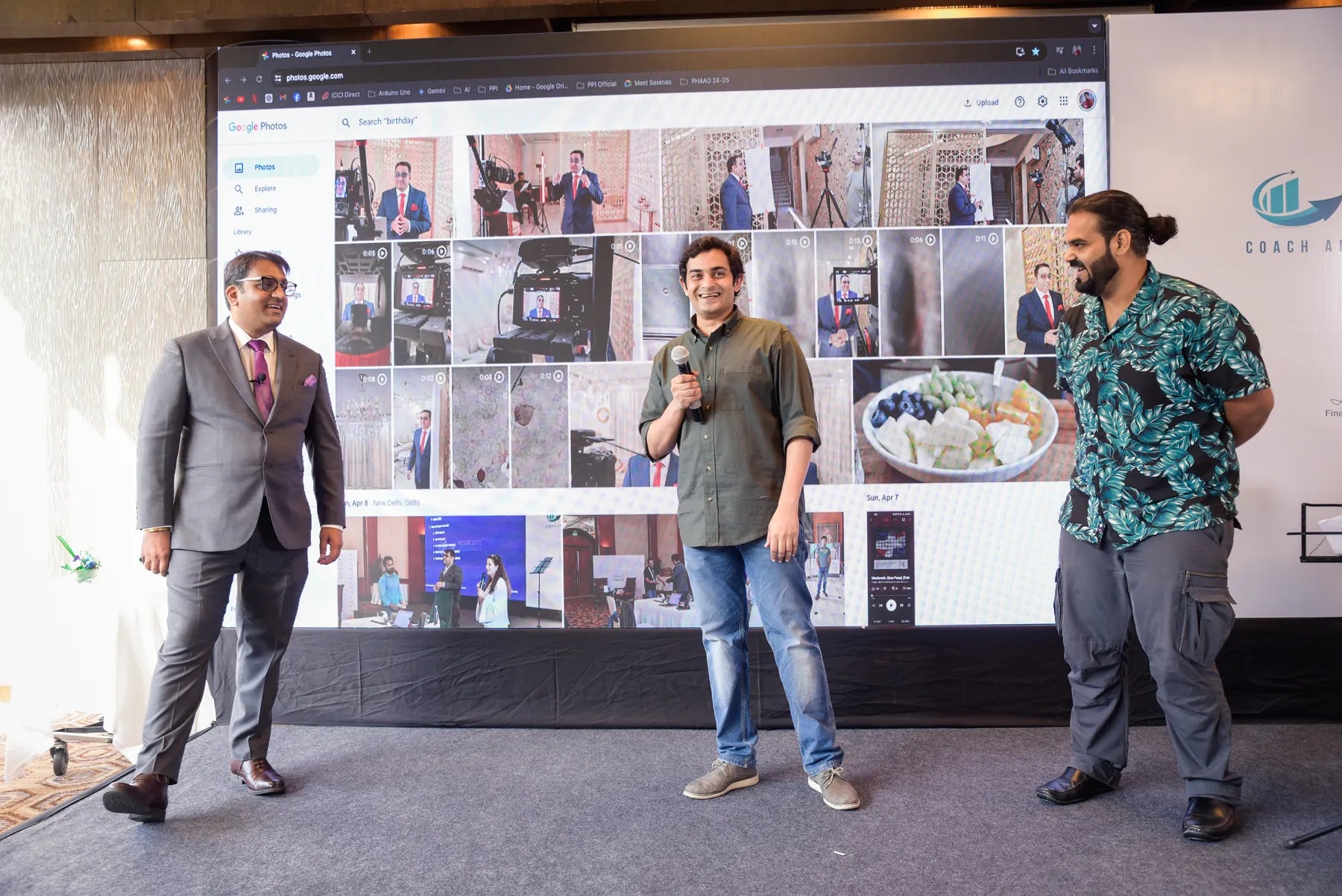 A speaker holding a microphone beside Anshul Kapoor and another guest in front of a screen at a programme event