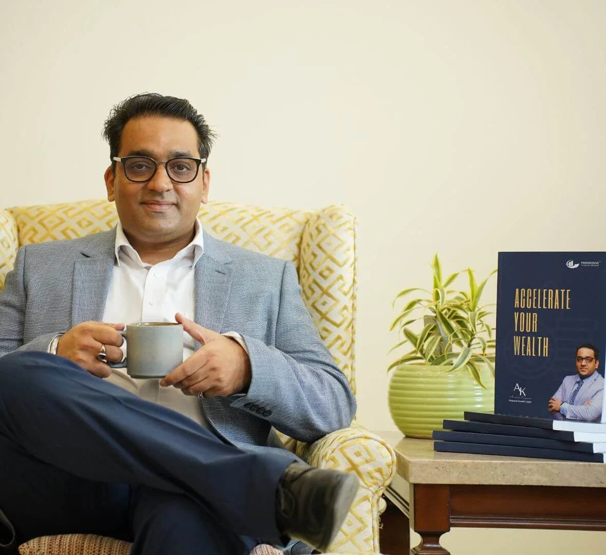 Anshul Kapoor seated in an armchair holding a cup, with a stack of books and a plant on a side table