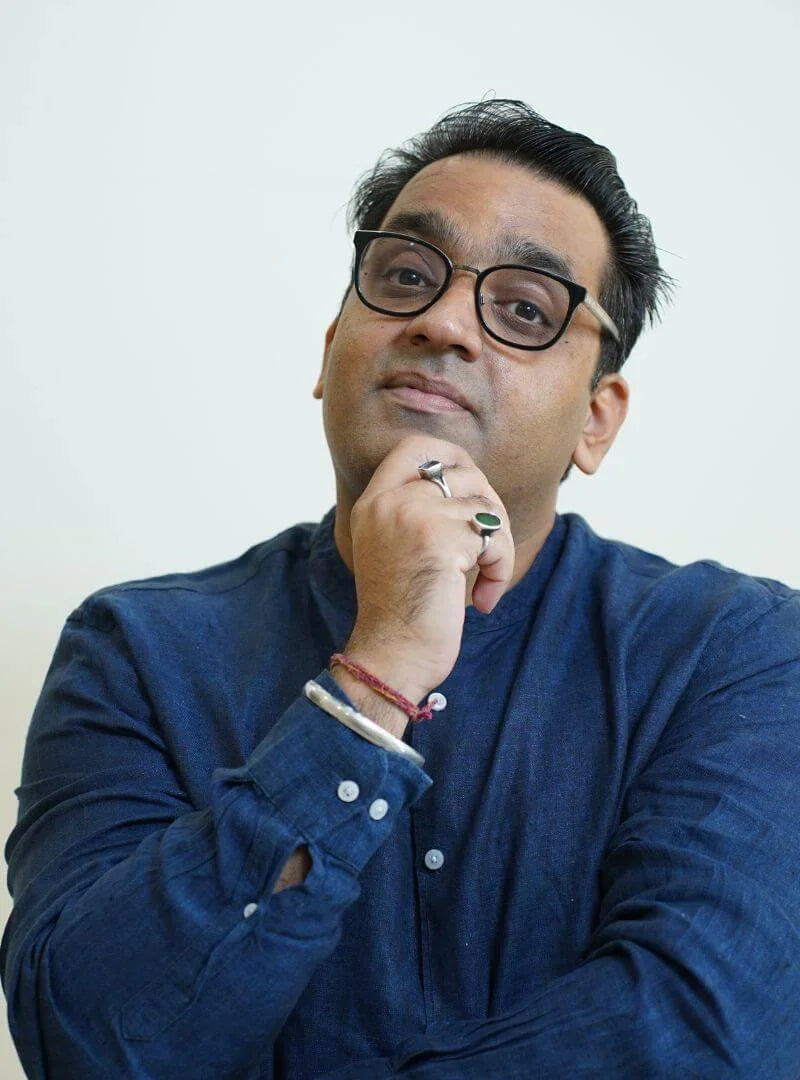 Anshul Kapoor with a hand on his chin, looking thoughtful, in a navy shirt