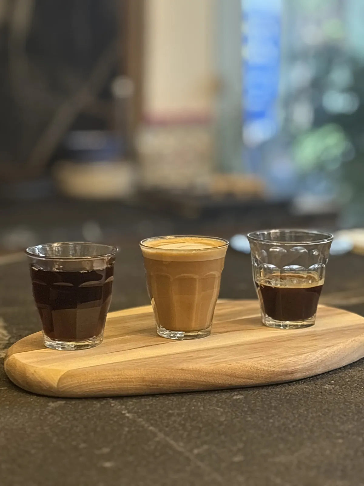 Three small glasses of coffee, from dark espresso to a milky coffee, on a round wooden paddle