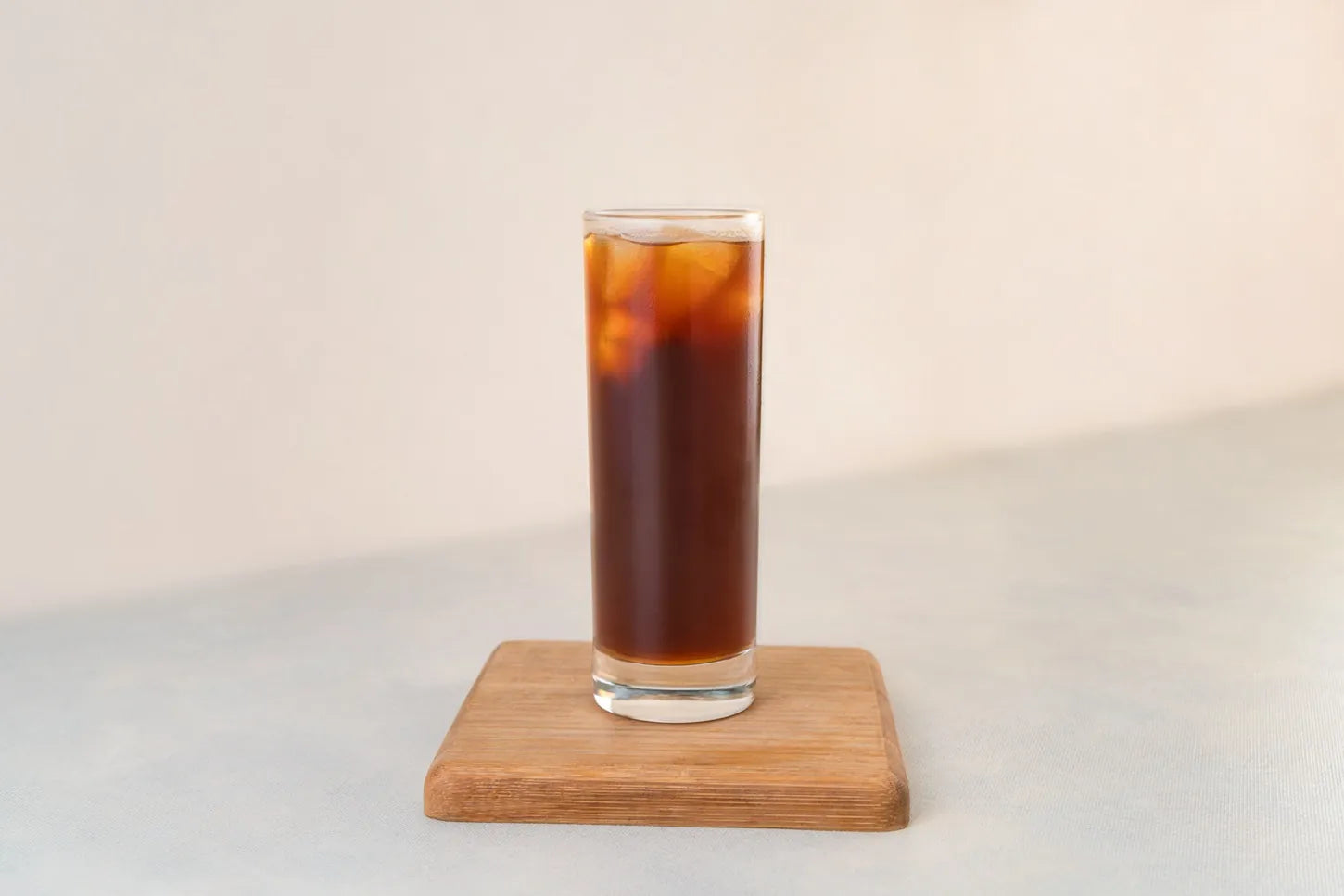 A tall glass of iced black coffee on a square wooden coaster against a pale background