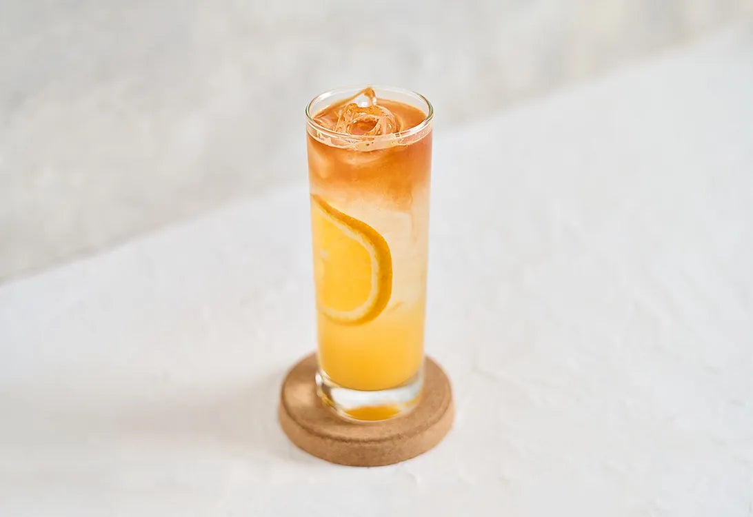A tall glass of orange-coloured iced drink with an orange slice, on a cork coaster