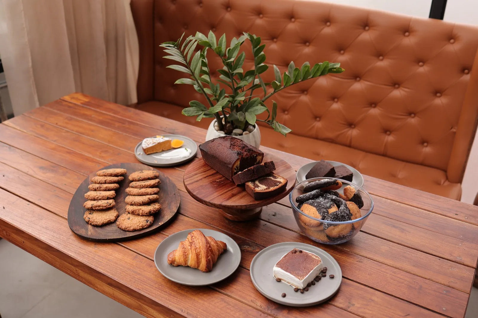 A wooden table in front of a tan leather sofa, laid with cookies, a loaf cake, a croissant and a dessert