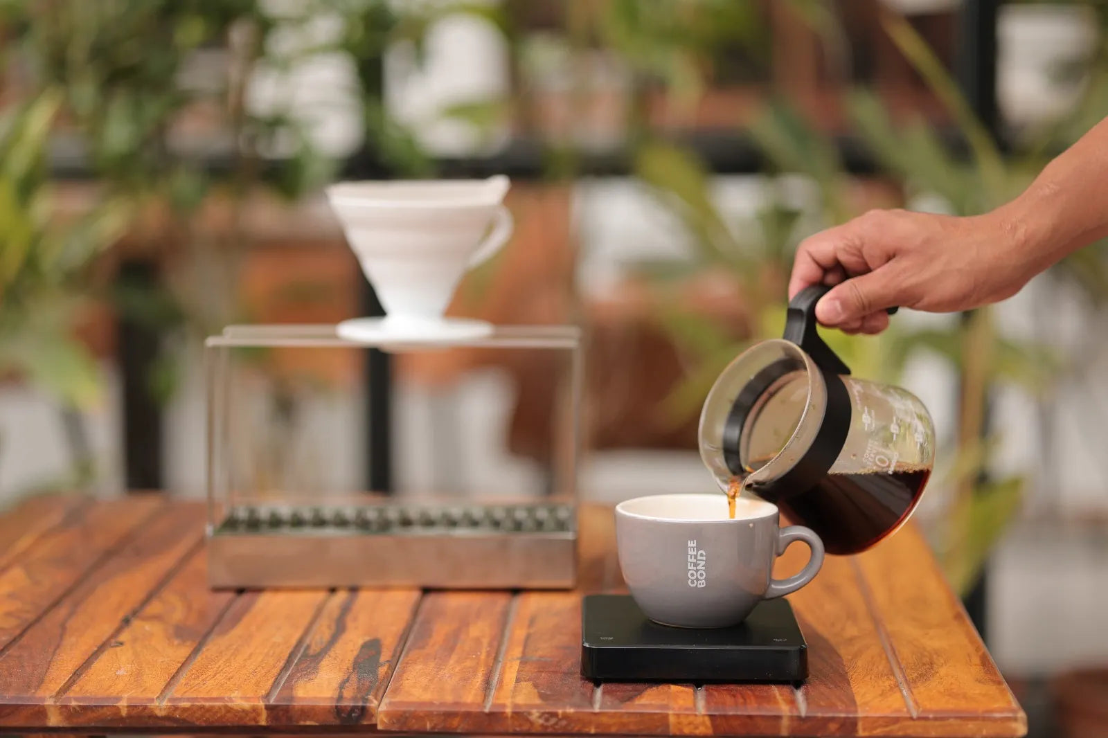 Coffee being poured from a glass server into a grey cup on a wooden table beside a pour-over stand