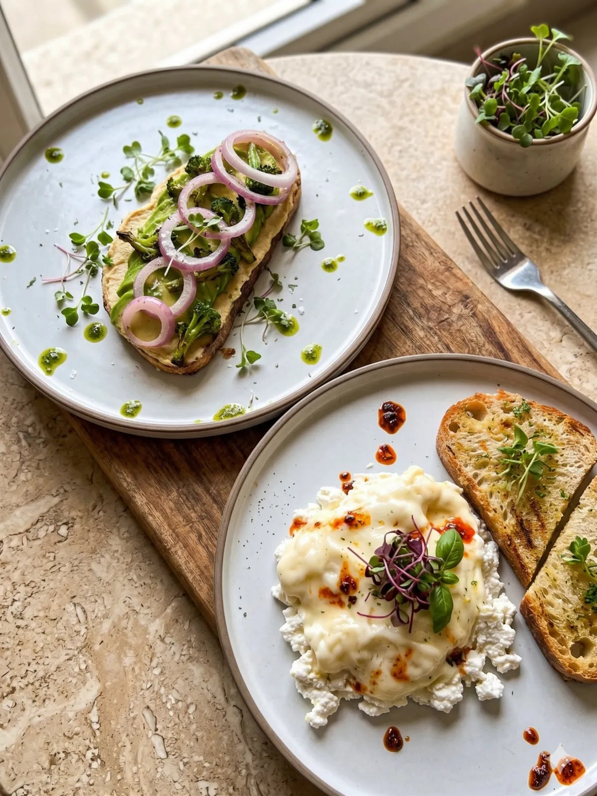 Two plates of toast with microgreens, one topped with avocado and pickled onion rings and one with ricotta and chilli oil, on a wooden board
