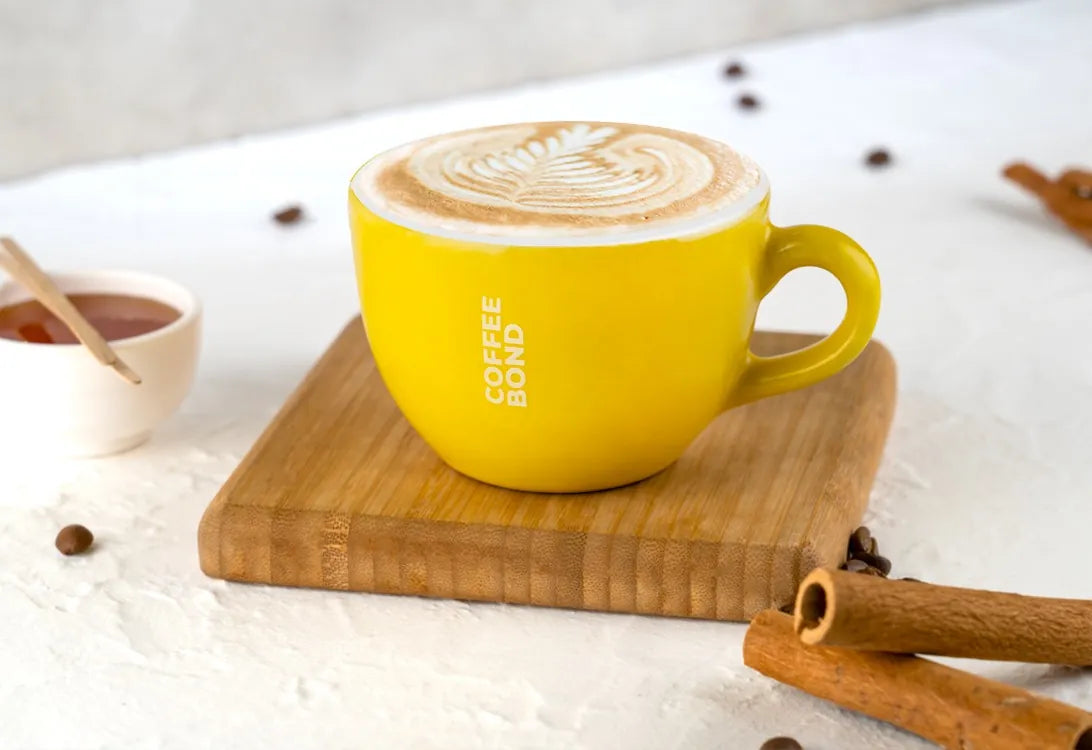 A latte with art in a yellow Coffee Bond cup on a wooden board, with cinnamon sticks
