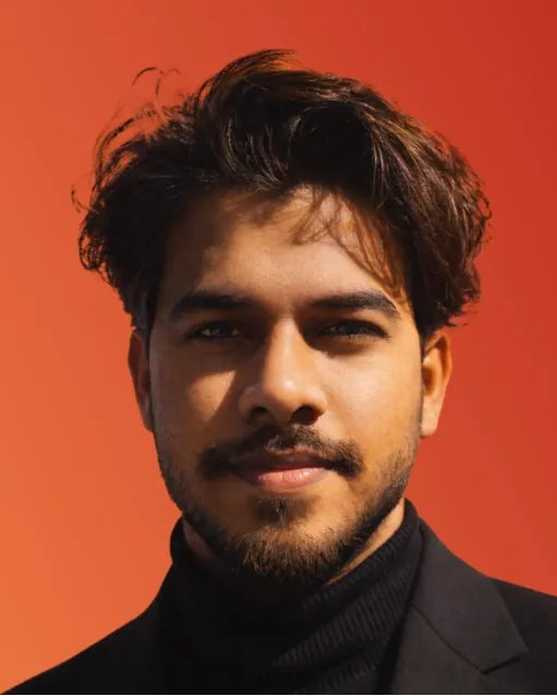 Portrait of Aarif Sheikh, Creative Head, in a dark jacket and roll-neck against an orange-red background